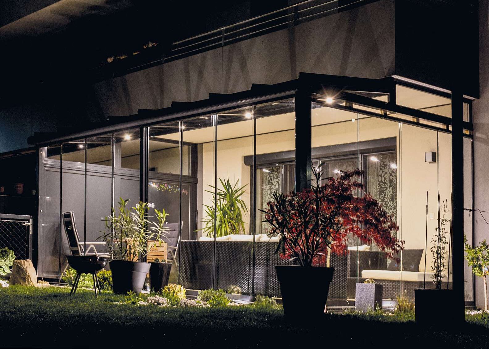 Eine mit Glaselementen geschlossene Terrassenüberdachung bei Nacht, von innen beleuchtet.
